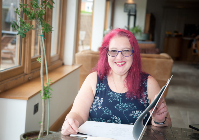 White woman Pollie Rafferty with pink hair sat at a desk and smiling at the camera